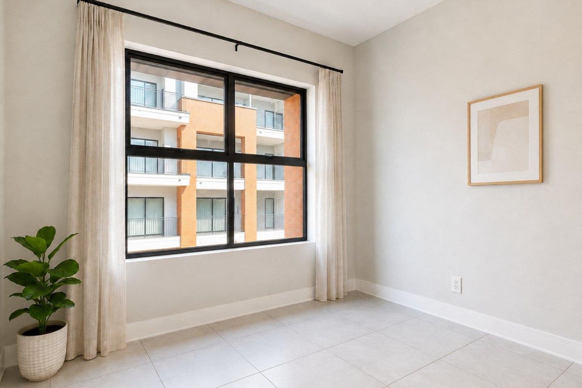 A bright, empty bedroom featuring neutral tiled flooring, clean walls, and a large black-framed window overlooking the apartment complex.