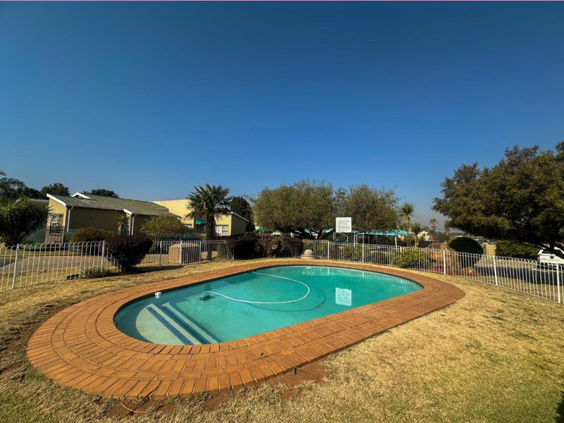 A communal swimming pool area featuring a brick-edged pool surrounded by a lawn and secure metal perimeter fencing.