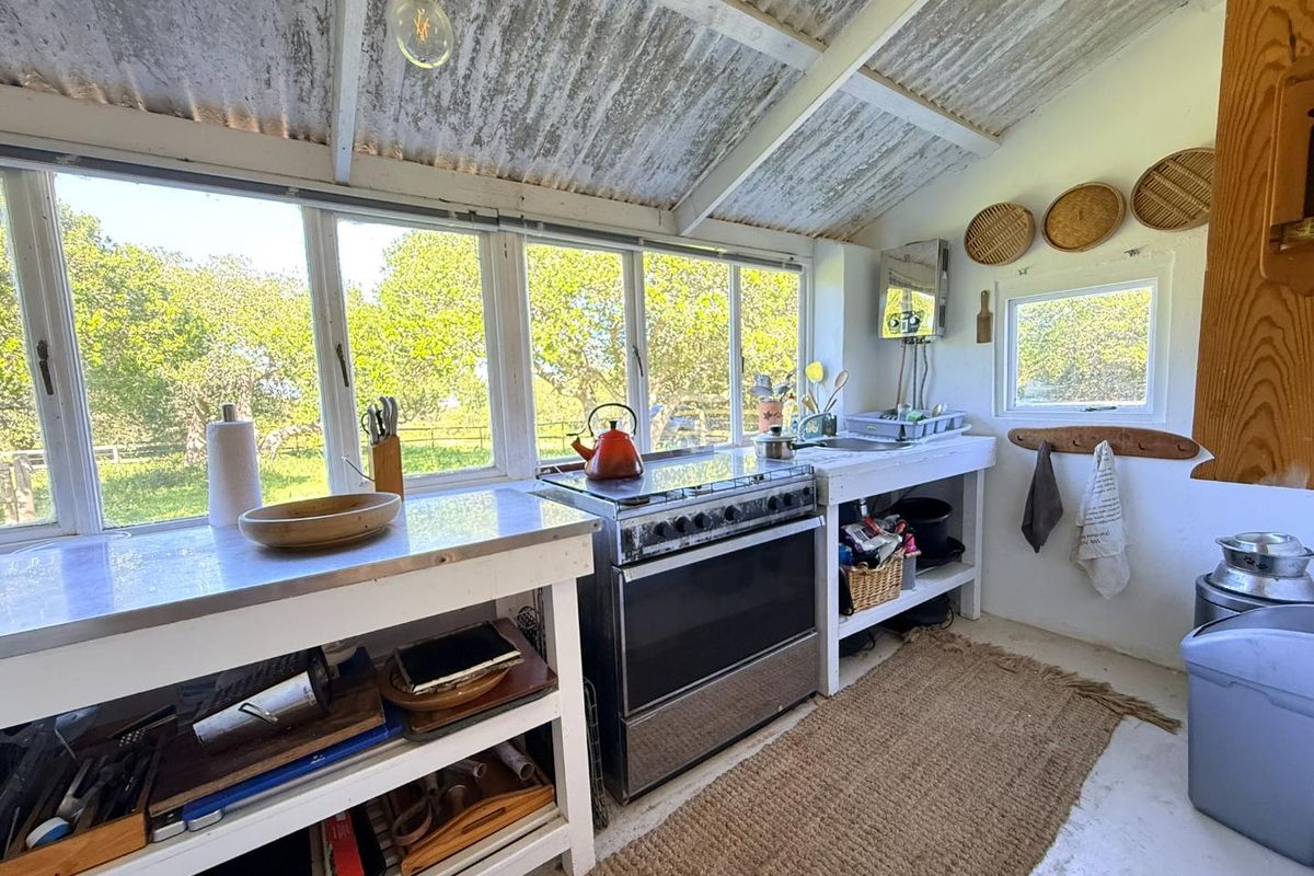 Rustic farm-style kitchen featuring a stainless steel workspace, freestanding oven, and large windows overlooking the orchard, with an exposed corrugated metal ceiling.