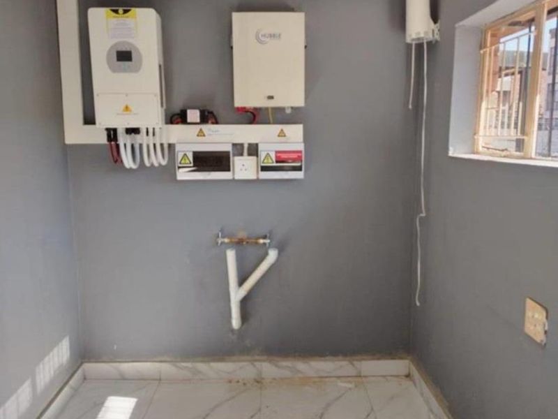 A clean utility space featuring a wall-mounted solar inverter, battery system, electrical distribution board, and plumbing connections for utility use.