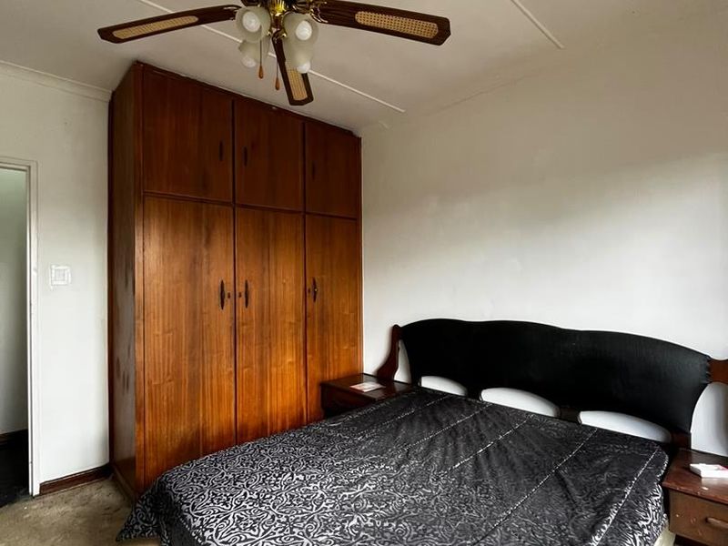 A bedroom featuring a built-in wooden wardrobe and a ceiling fan, with carpet flooring and visible wear on the ceiling panels.