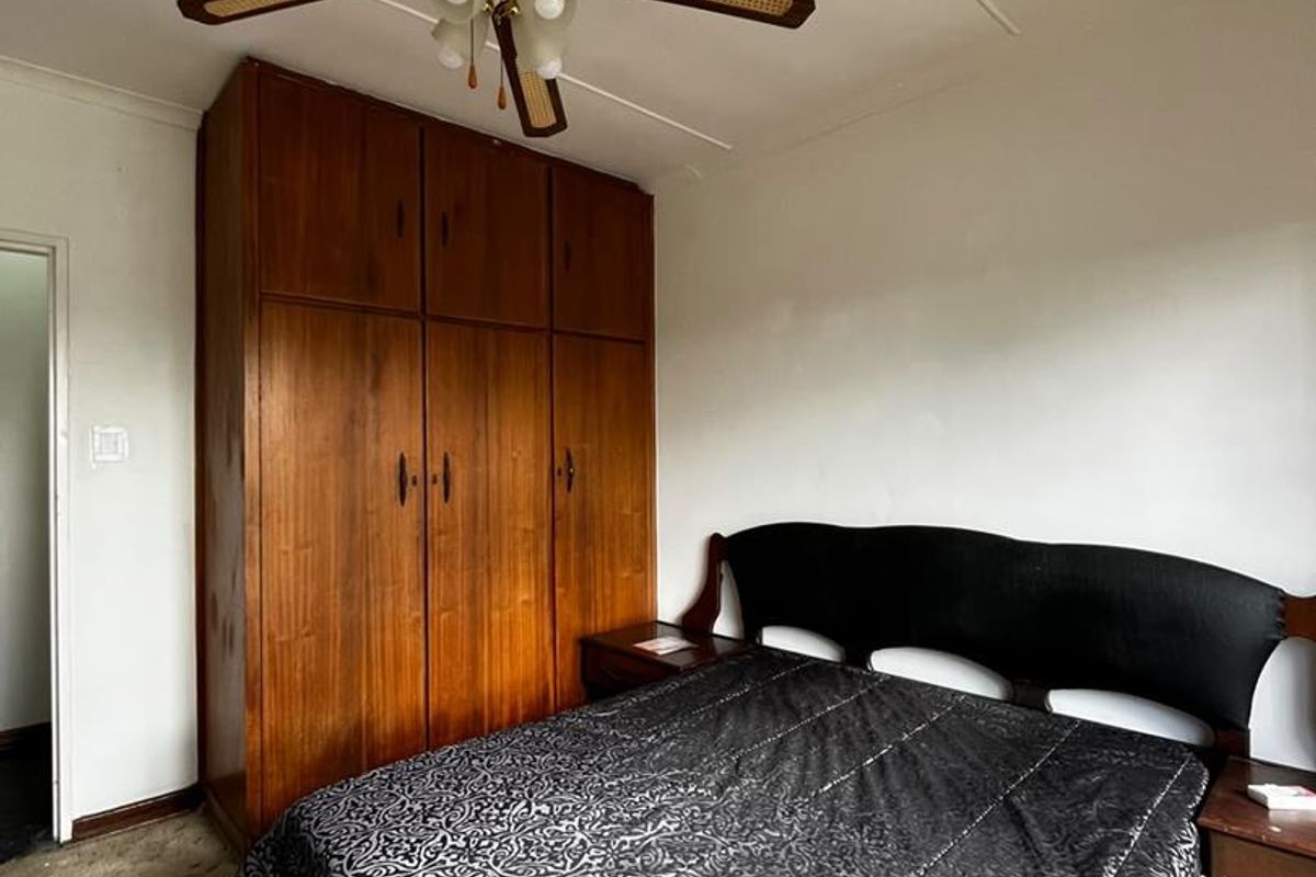 A bedroom featuring a built-in wooden wardrobe and a ceiling fan, with carpet flooring and visible wear on the ceiling panels.