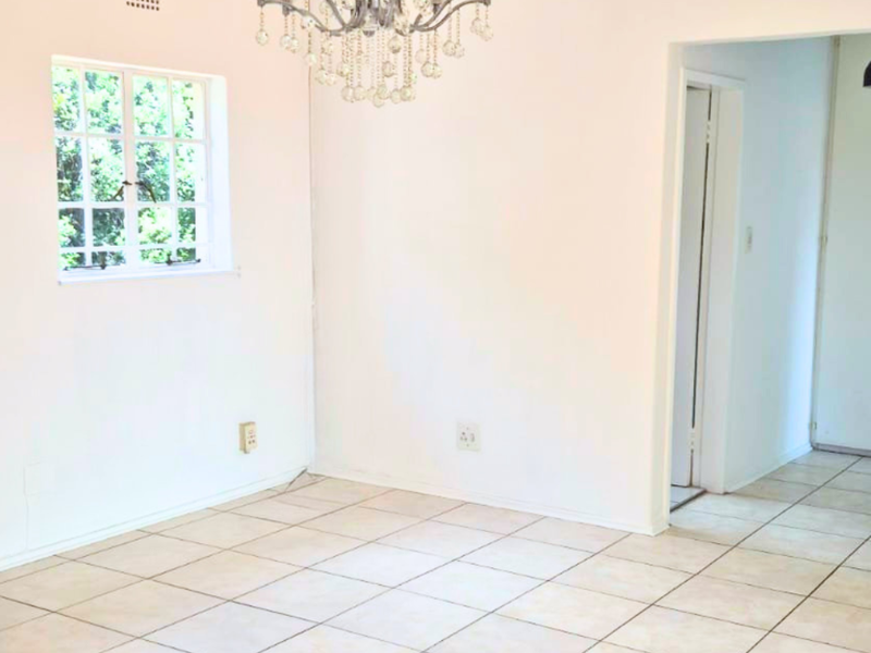 A bright, empty dining area featuring neutral ceramic tile flooring, a decorative crystal chandelier, and a classic casement window.