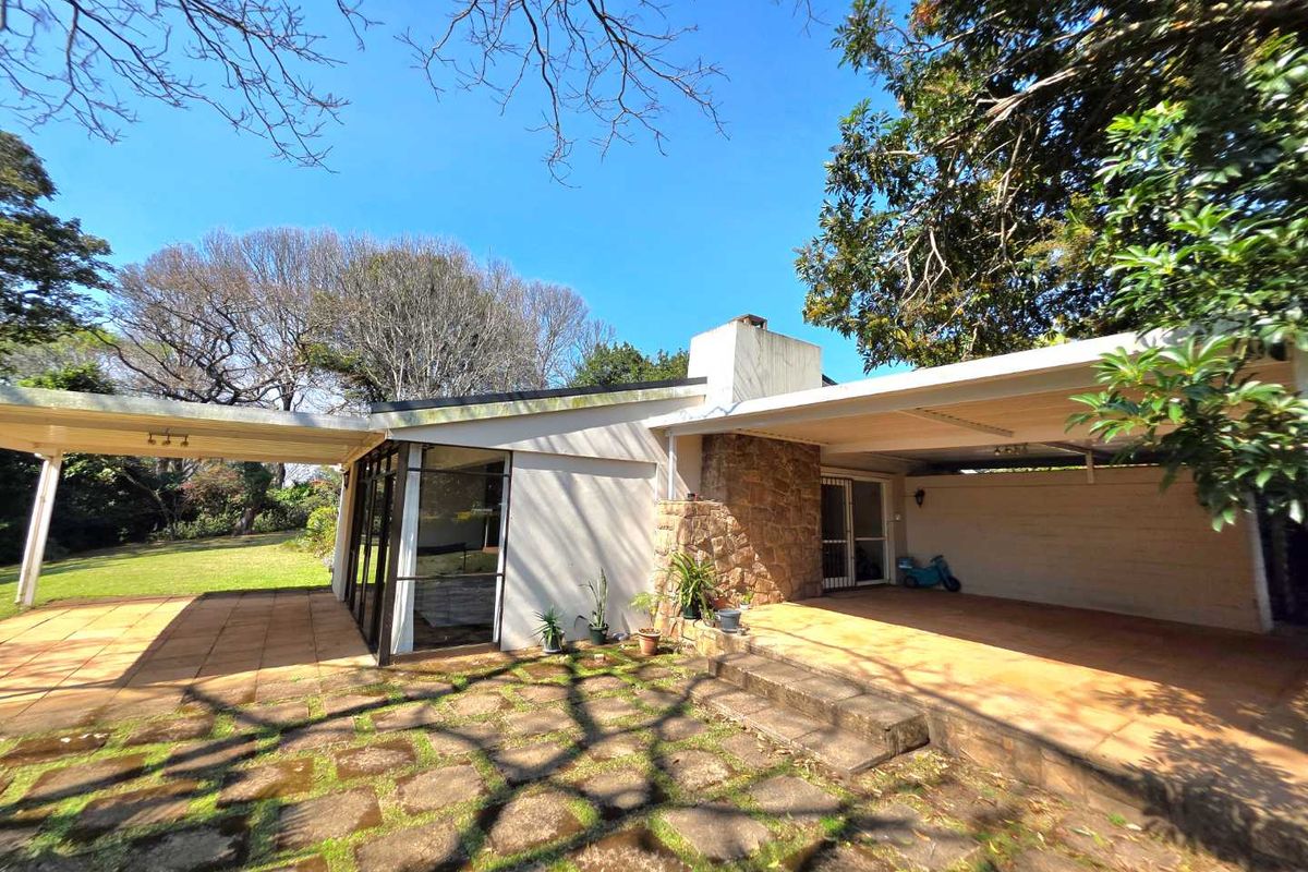 Rear view of the property featuring a covered patio, stone chimney, and paved outdoor area surrounded by mature trees.