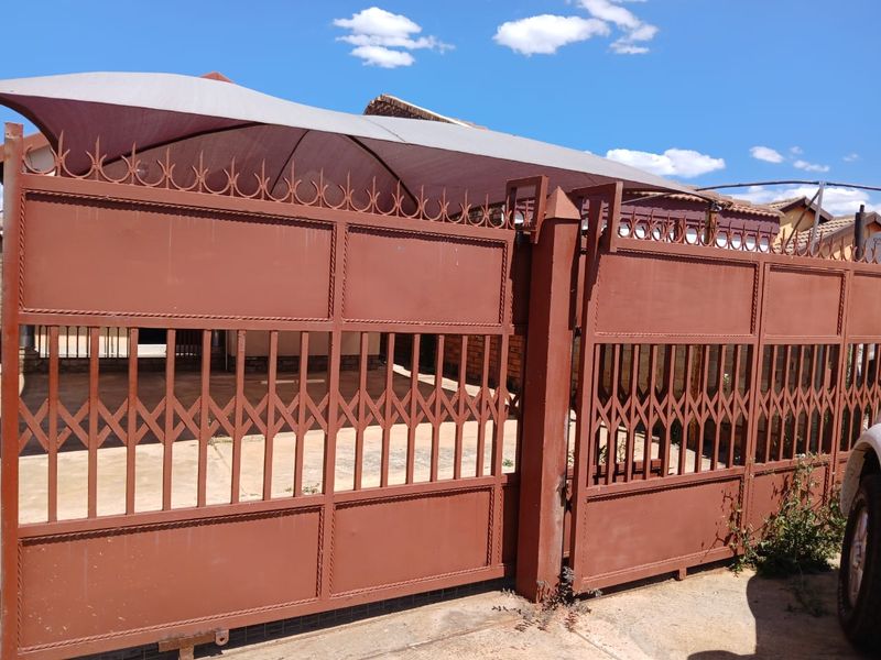 Front view of the property featuring a large metal security gate, paved driveway area, and a protective shade sail structure.