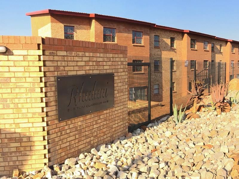 Exterior view of the apartment complex entrance featuring a brick boundary wall, metal security fencing, and landscaped rock garden.
