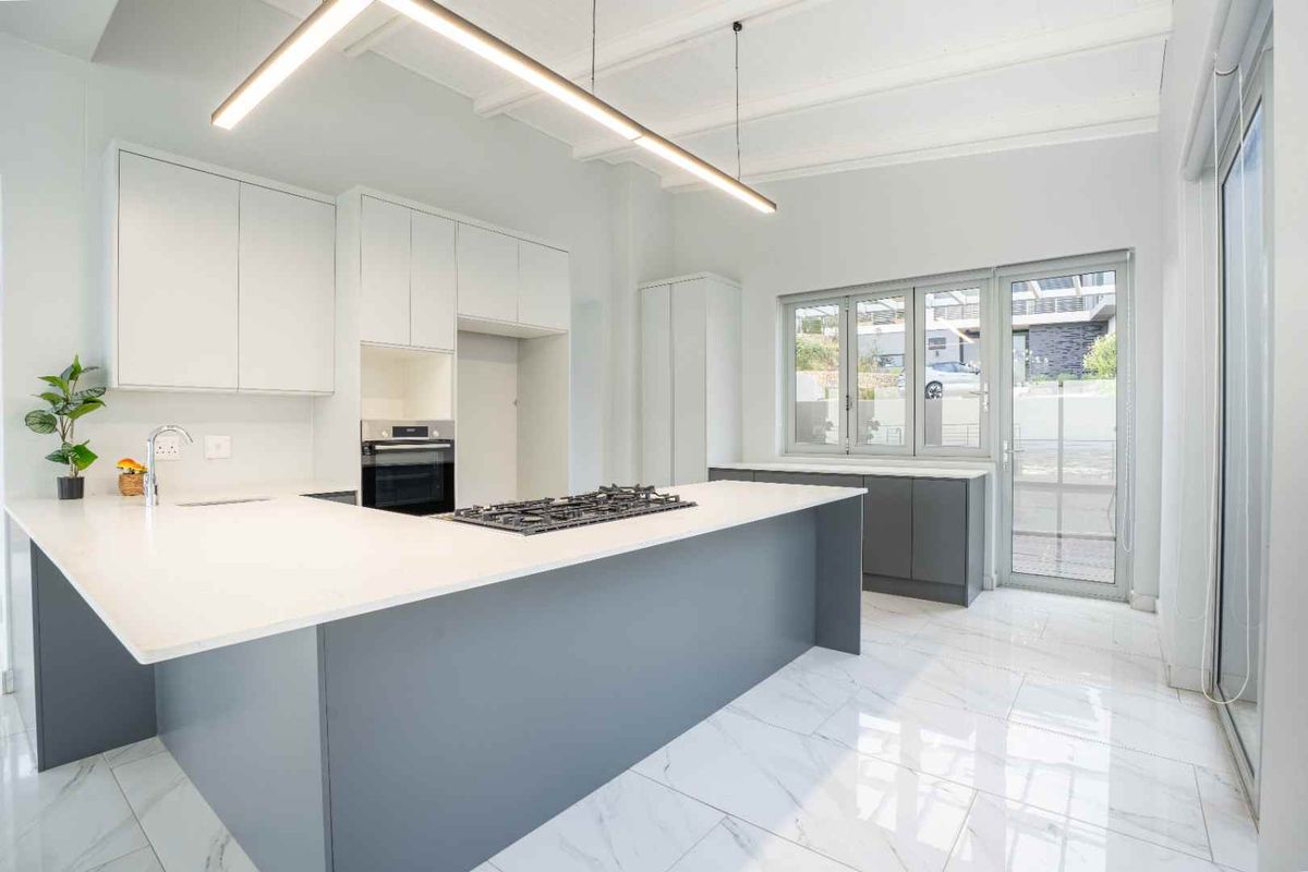 Modern kitchen featuring sleek grey and white cabinetry, stone countertops, a gas hob, built-in oven, and elegant marble-look flooring with ample natural light.
