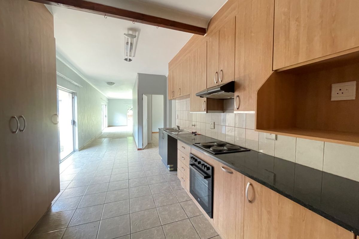 A functional kitchen featuring light wood cabinetry, dark countertops, built-in oven and hob, and tiled flooring, leading into an open-plan living area.