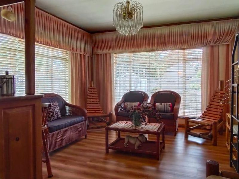 A living room featuring laminate flooring, large windows with venetian blinds and pelmet curtains, and a central crystal chandelier.