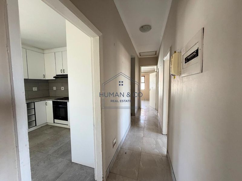 A view of a hallway leading to other rooms, with a glimpse into an adjacent kitchen featuring white cabinetry and tiled flooring.