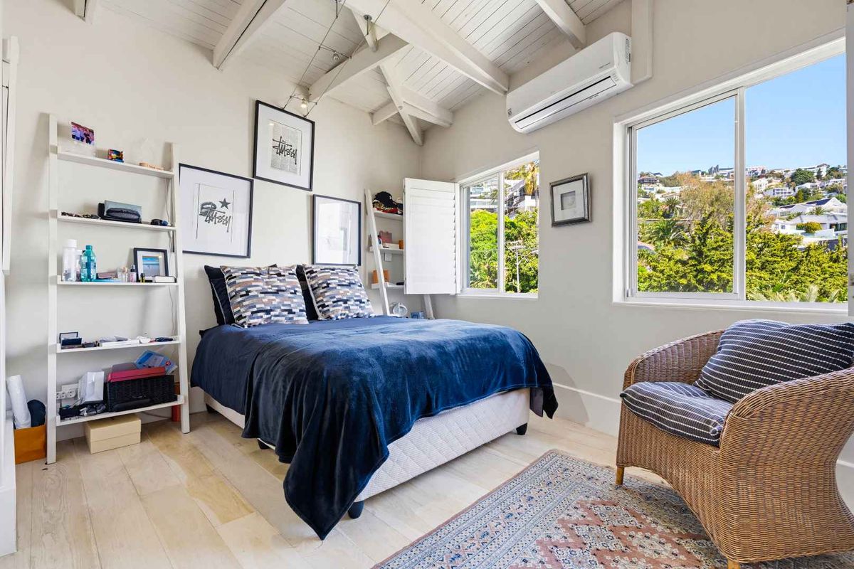 A bright bedroom featuring exposed timber ceiling beams, light timber flooring, a split-system air conditioner, and large windows with plantation shutters.