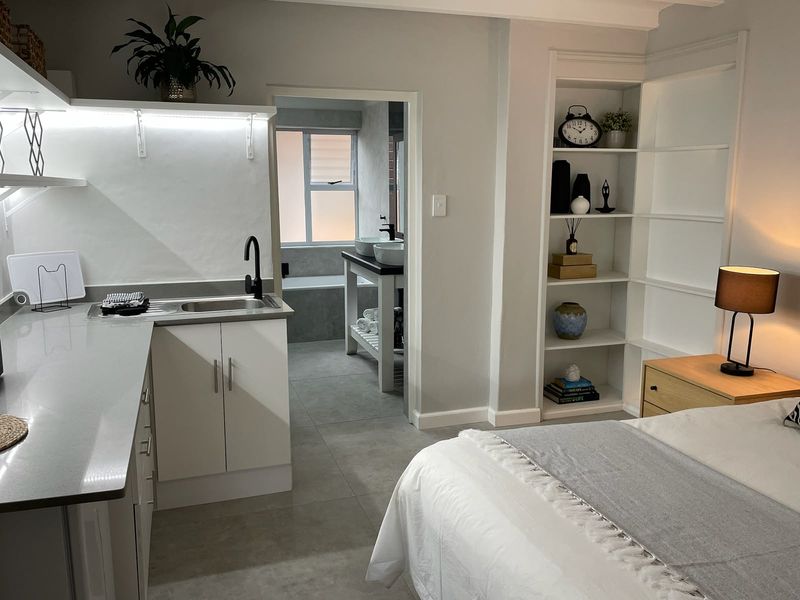 A modern bedroom featuring built-in shelving, a compact kitchenette area with stone countertops, and direct access to an en-suite bathroom with tiled finishes.