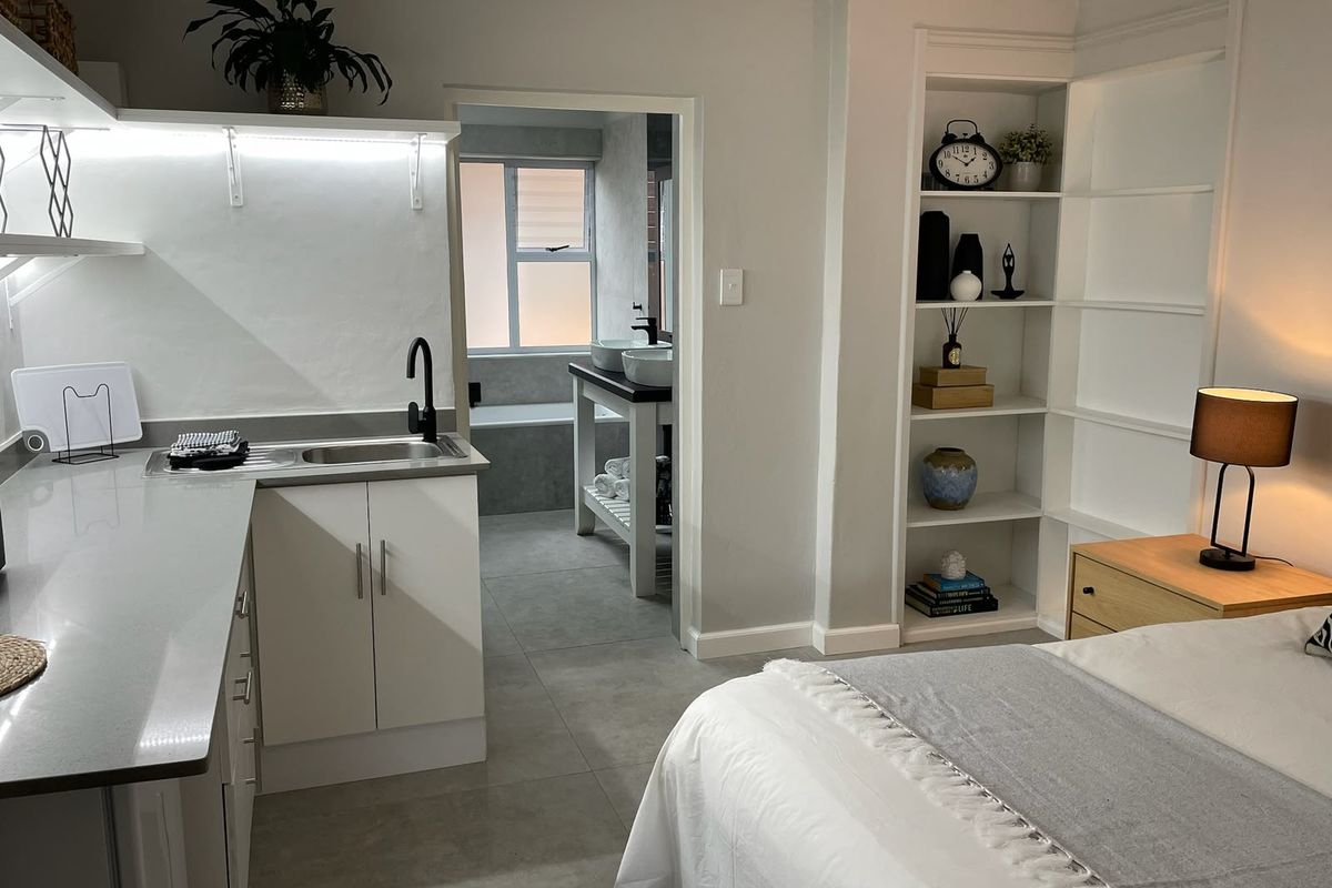 A modern bedroom featuring built-in shelving, a compact kitchenette area with stone countertops, and direct access to an en-suite bathroom with tiled finishes.