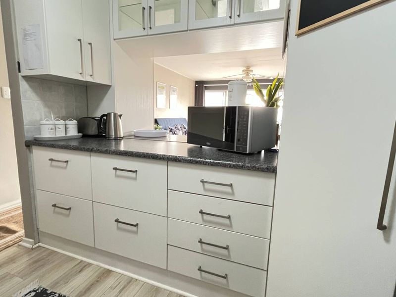 A clean, modern kitchen featuring white cabinetry, dark laminate countertops, and integrated microwave space, with a convenient pass-through window to the living area.