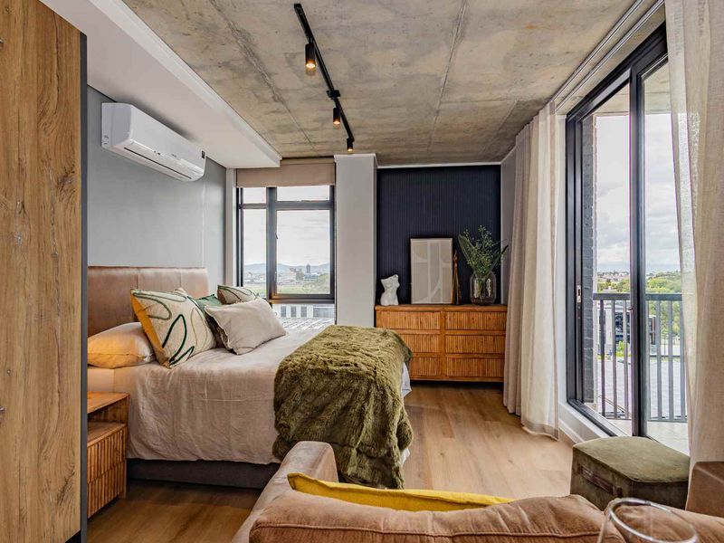 Modern bedroom featuring an exposed concrete ceiling, timber flooring, split-system air conditioning, and large sliding glass doors opening to a balcony.