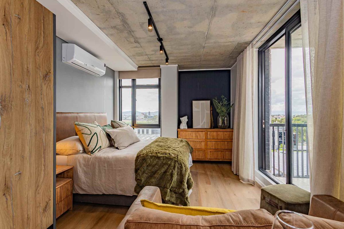 Modern bedroom featuring an exposed concrete ceiling, timber flooring, split-system air conditioning, and large sliding glass doors opening to a balcony.