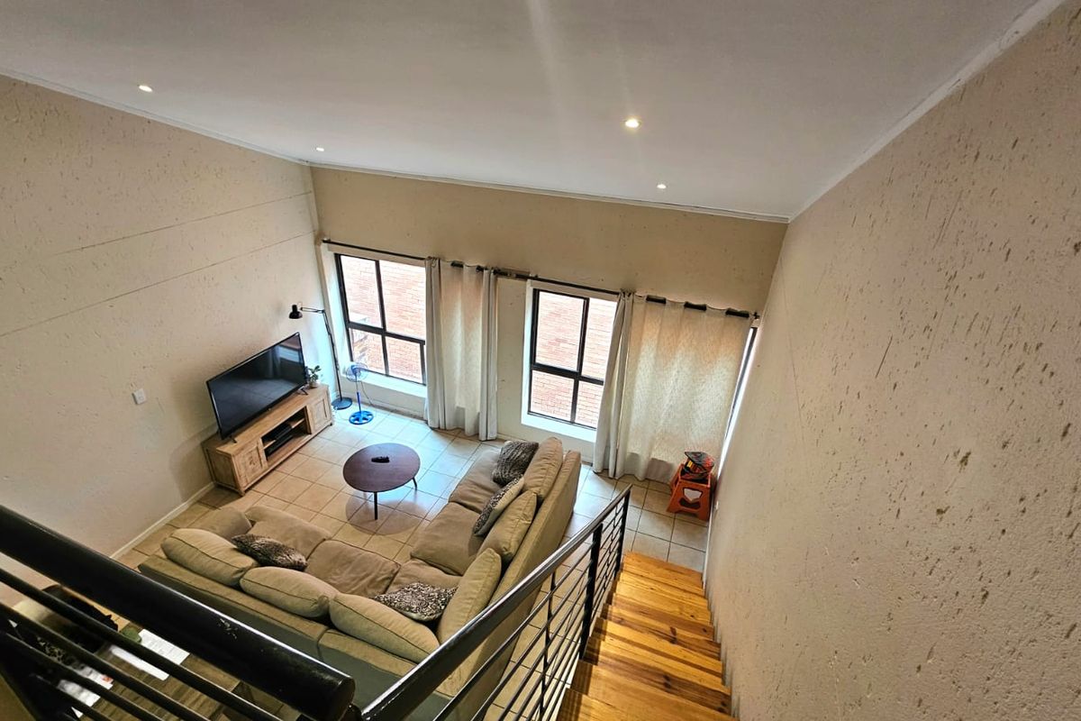 Spacious double-volume living area featuring tiled flooring, large windows for natural light, and a staircase with a metal balustrade.