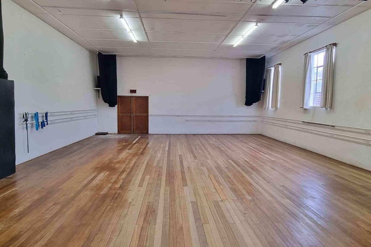 A large, open-plan studio space featuring polished hardwood flooring, wall-mounted ballet barres, and functional overhead fluorescent lighting.