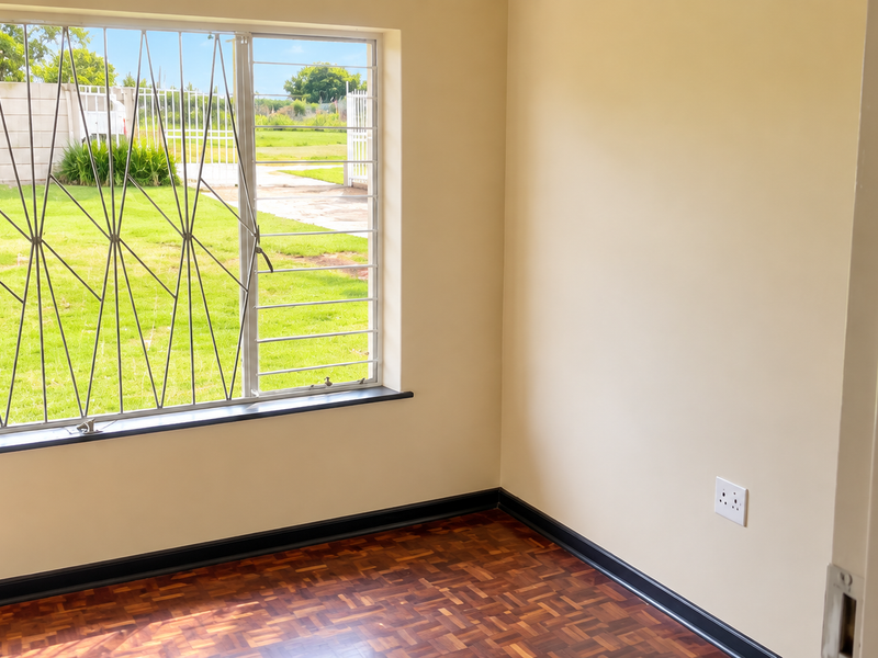 A bright, empty bedroom featuring polished parquet flooring, neutral walls, and a large window fitted with security bars overlooking the garden.