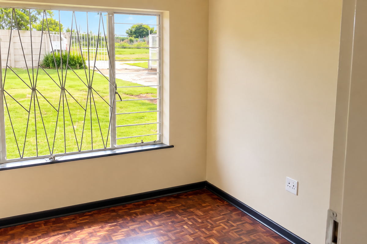 A bright, empty bedroom featuring polished parquet flooring, neutral walls, and a large window fitted with security bars overlooking the garden.