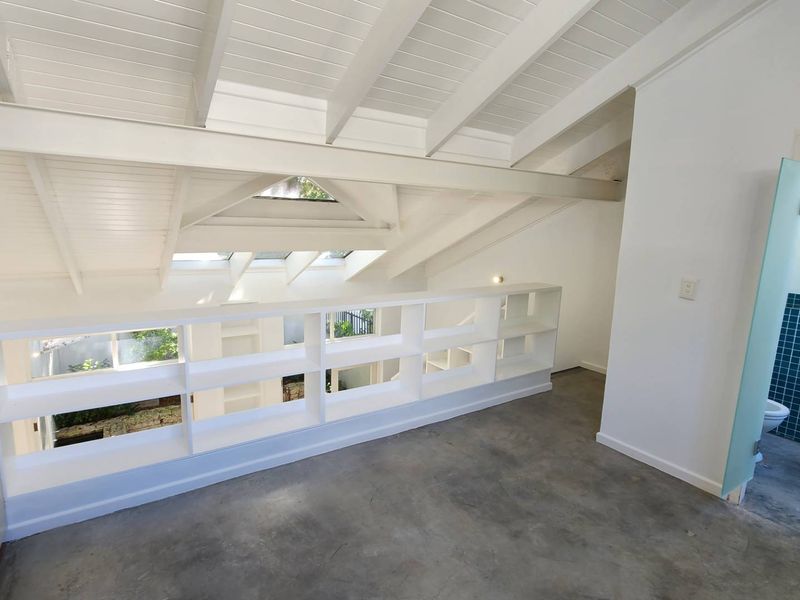 A bright, open-plan loft bedroom featuring exposed white timber beams, skylights, concrete flooring, and a built-in shelving unit overlooking the lower level.