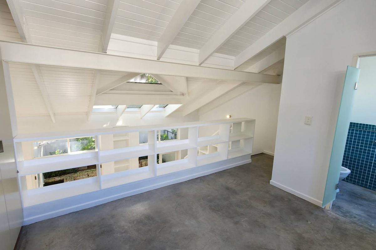 A bright, open-plan loft bedroom featuring exposed white timber beams, skylights, concrete flooring, and a built-in shelving unit overlooking the lower level.