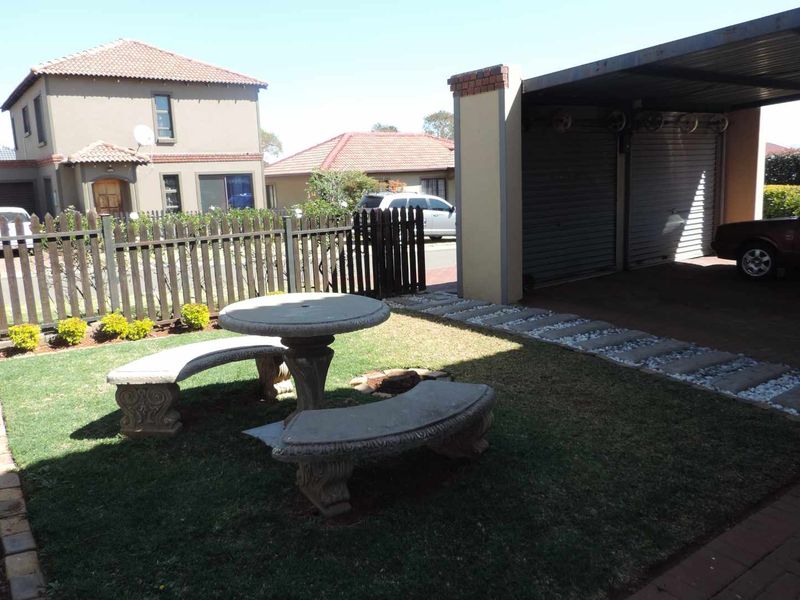 A view of the rear garden area featuring a carport, paved walkway, and a picket fence separating the property from the street.
