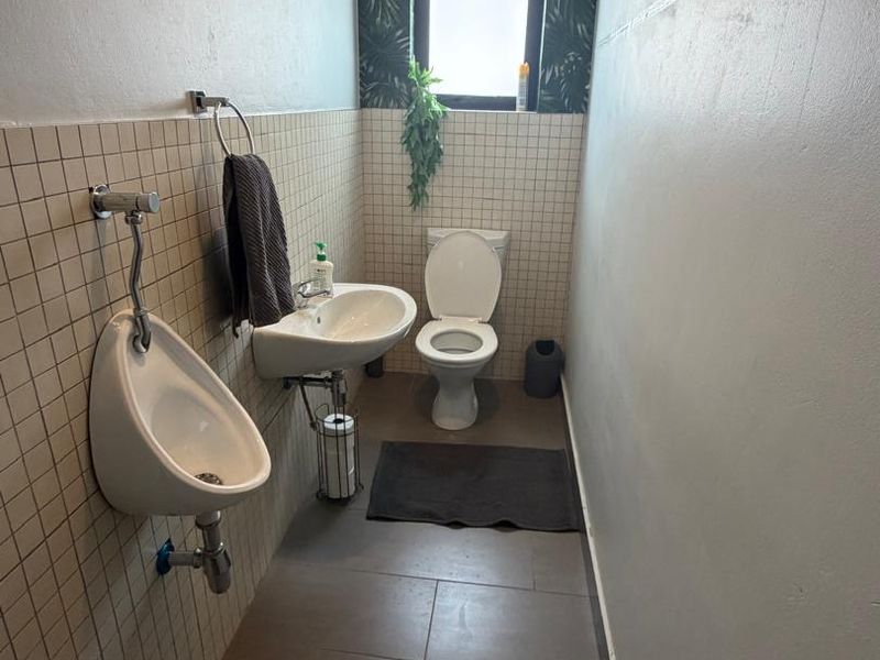 A functional commercial bathroom featuring a wall-mounted urinal, ceramic washbasin, and flush toilet with tiled finishes and a window for natural light.