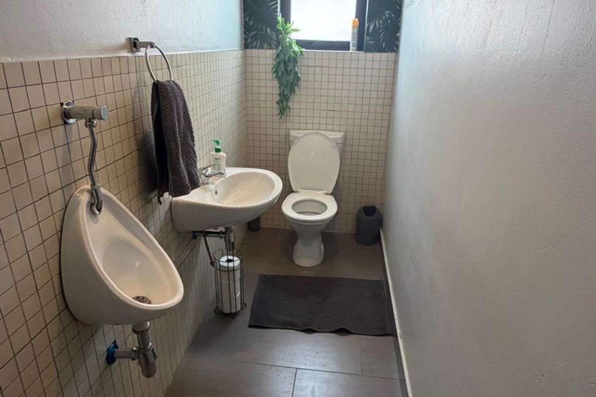 A functional commercial bathroom featuring a wall-mounted urinal, ceramic washbasin, and flush toilet with tiled finishes and a window for natural light.