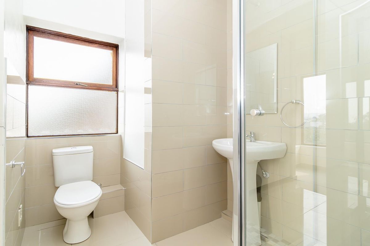 A clean, functional bathroom featuring a pedestal washbasin, a toilet, and a glass-enclosed shower with neutral wall and floor tiling.