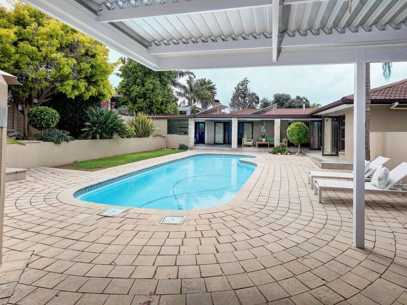 A well-maintained outdoor pool area featuring a paved deck, covered patio space, and a built-in fireplace, perfect for outdoor entertaining.