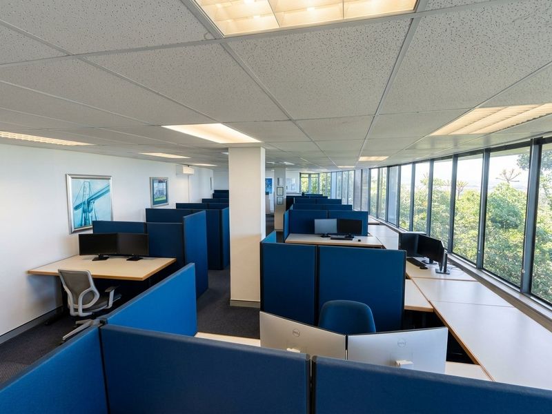 Open-plan office space featuring a suspended ceiling, large windows with natural light, and a functional layout with built-in workstation partitions.