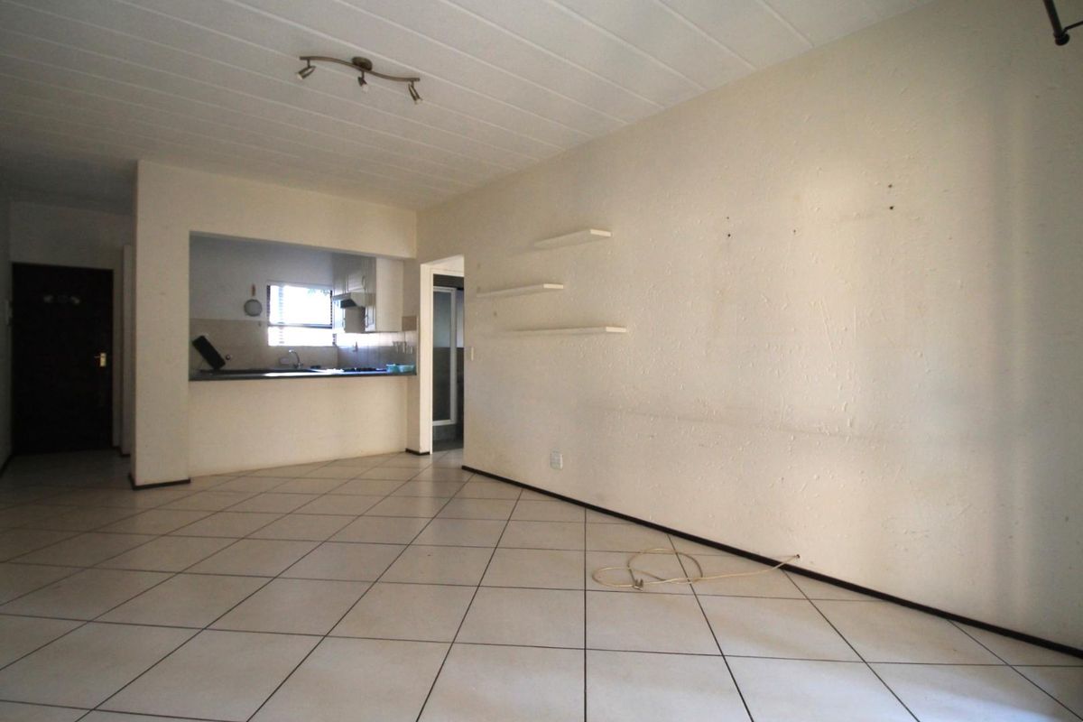 An open-plan living area featuring tiled flooring and a pass-through kitchen window, requiring cosmetic wall repairs and a fresh coat of paint.