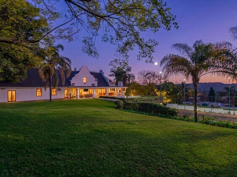 A beautiful rear view of a Cape Dutch style home at dusk, featuring a spacious lawn, covered patio, and a private tennis court.