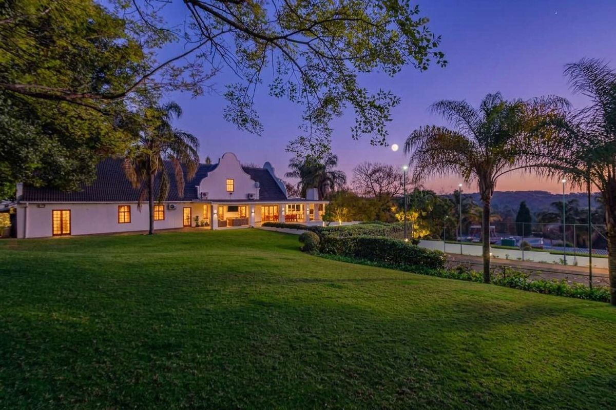 A beautiful rear view of a Cape Dutch style home at dusk, featuring a spacious lawn, covered patio, and a private tennis court.