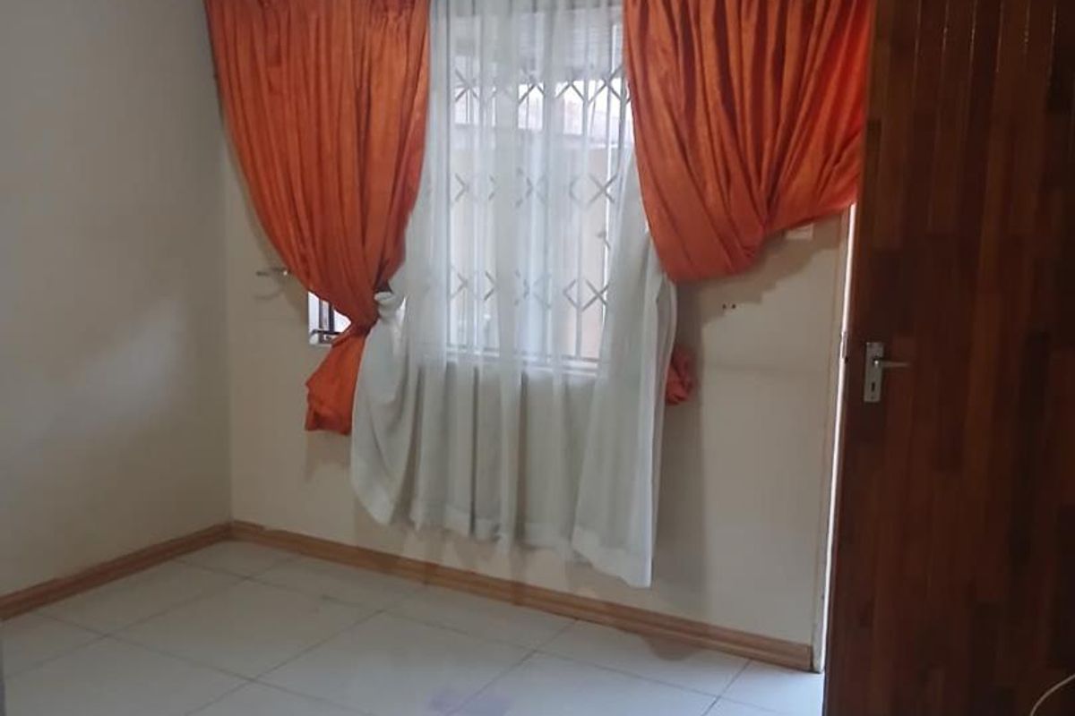 A simple, empty bedroom featuring tiled flooring, a window with security bars, and a wooden door.