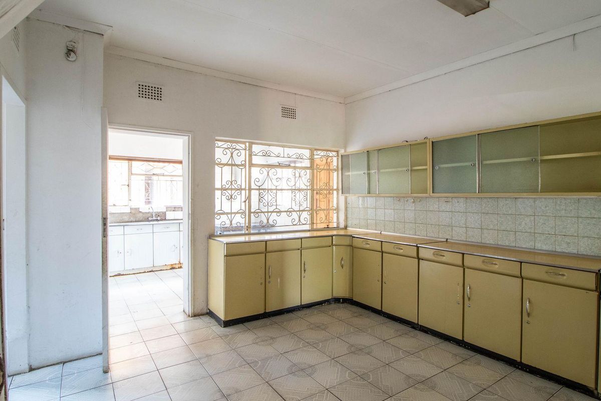 A dated kitchen featuring original cabinetry, tiled flooring, and a view into an adjacent scullery area with security bars on the windows.