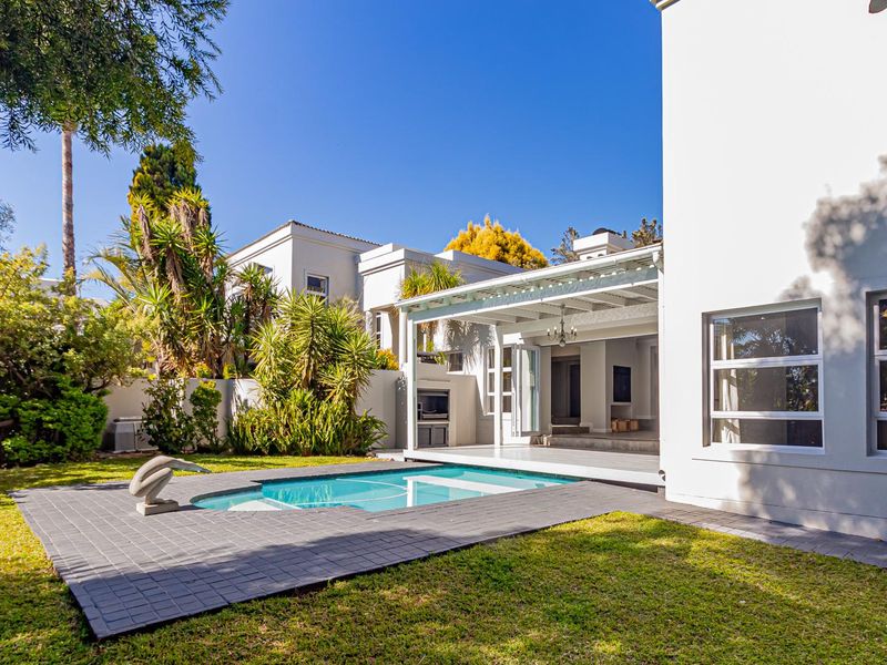 A well-maintained outdoor entertainment area featuring a swimming pool, paved deck, and a covered patio with a built-in braai, set within a landscaped garden.