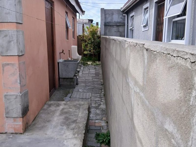 A narrow side passage between two buildings featuring a concrete wash trough, paved walkway, and boundary wall.