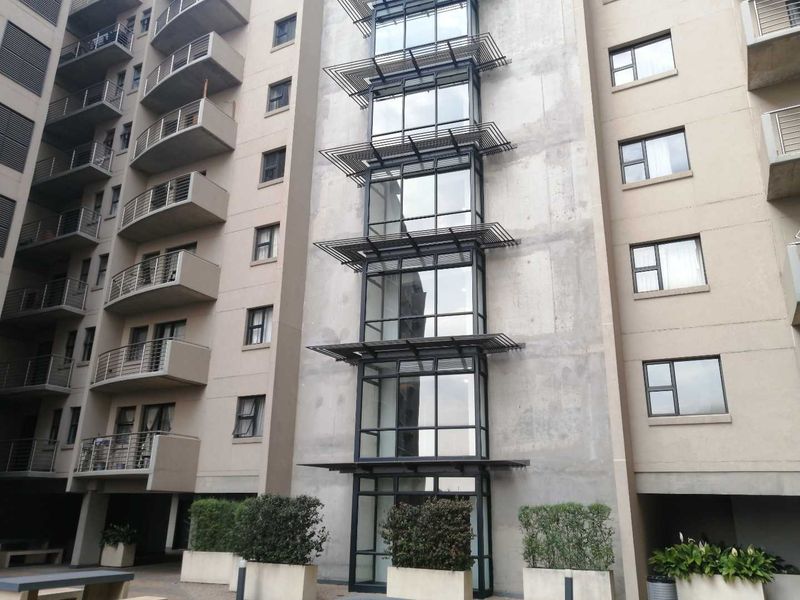 Exterior view of a modern apartment building featuring a concrete facade, metal balcony railings, and a prominent vertical glass curtain wall with sun louvres.