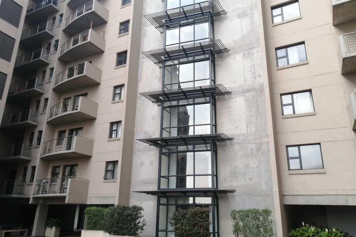 Exterior view of a modern apartment building featuring a concrete facade, metal balcony railings, and a prominent vertical glass curtain wall with sun louvres.