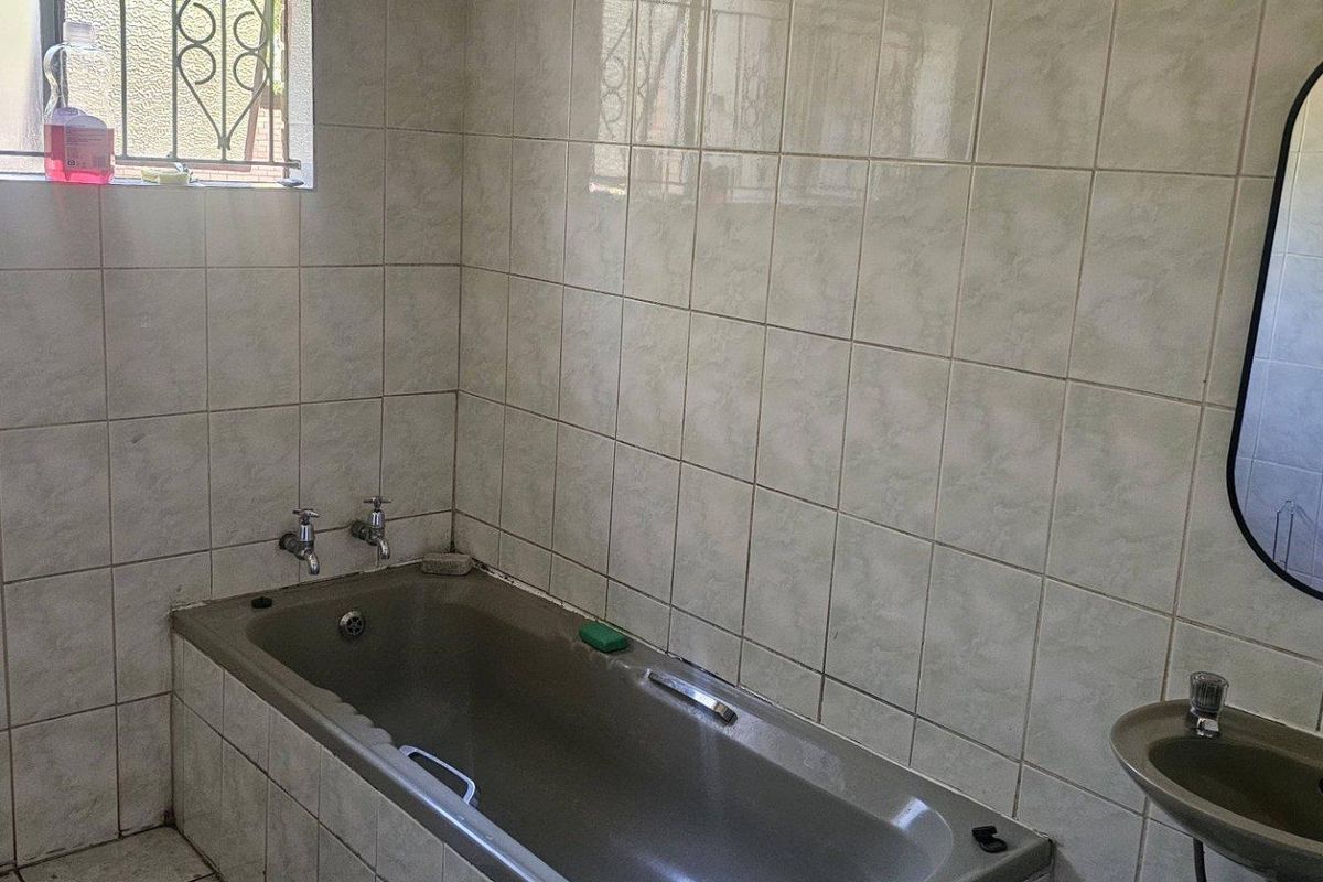 A dated bathroom featuring a built-in bathtub, wall-mounted basin, and full-height wall tiling with security bars on the window.