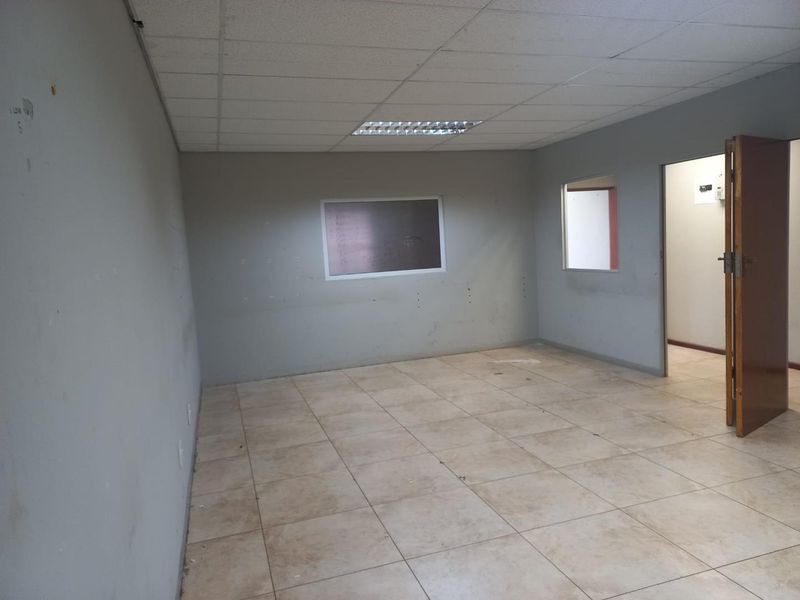 An empty office space featuring tiled flooring, a suspended ceiling with recessed lighting, and an internal window looking into an adjacent room.