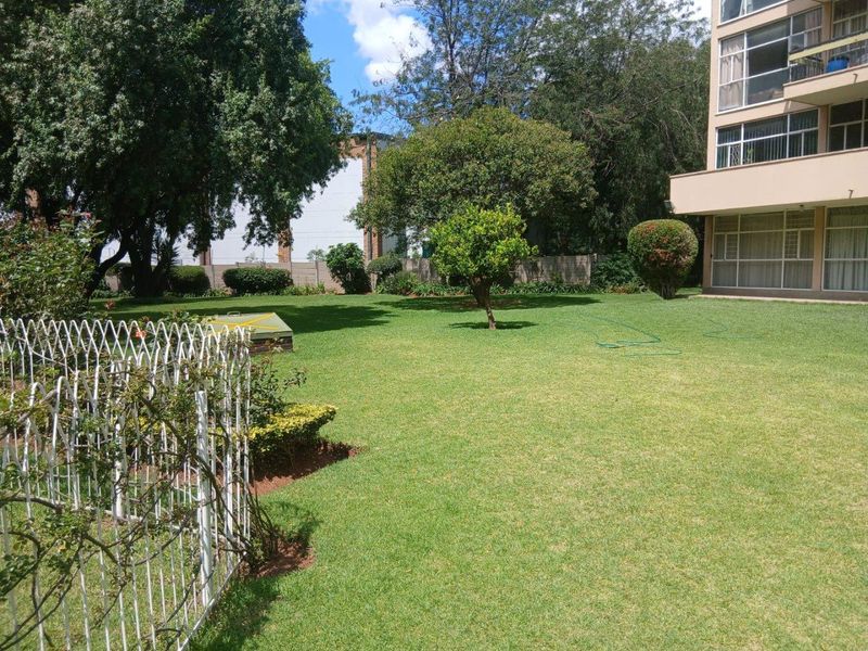A well-maintained communal garden area featuring a lush green lawn, mature trees, and a secure perimeter wall, adjacent to the apartment building.