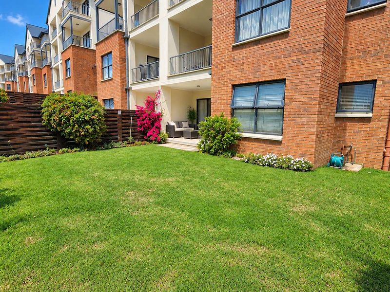 A well-maintained rear garden area featuring a manicured lawn, brick apartment facade, and a private covered patio space.