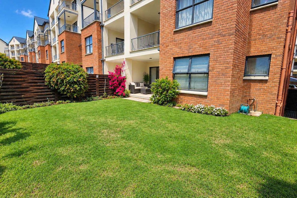 A well-maintained rear garden area featuring a manicured lawn, brick apartment facade, and a private covered patio space.