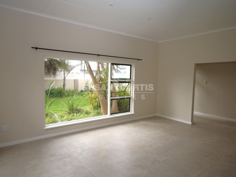 A bright, empty living room featuring neutral tiled flooring, recessed lighting, and a large window overlooking the garden.