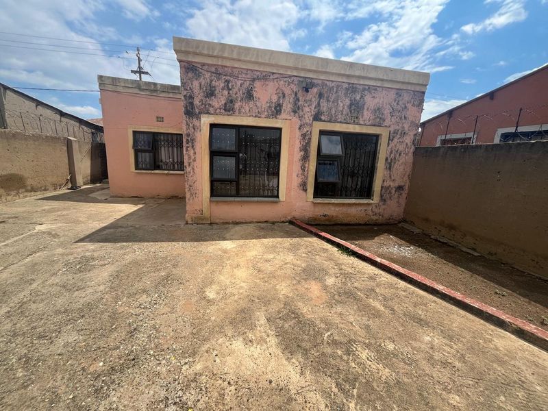 Rear exterior view of the property featuring a paved yard, boundary walls with security fencing, and a building facade requiring maintenance and repainting.