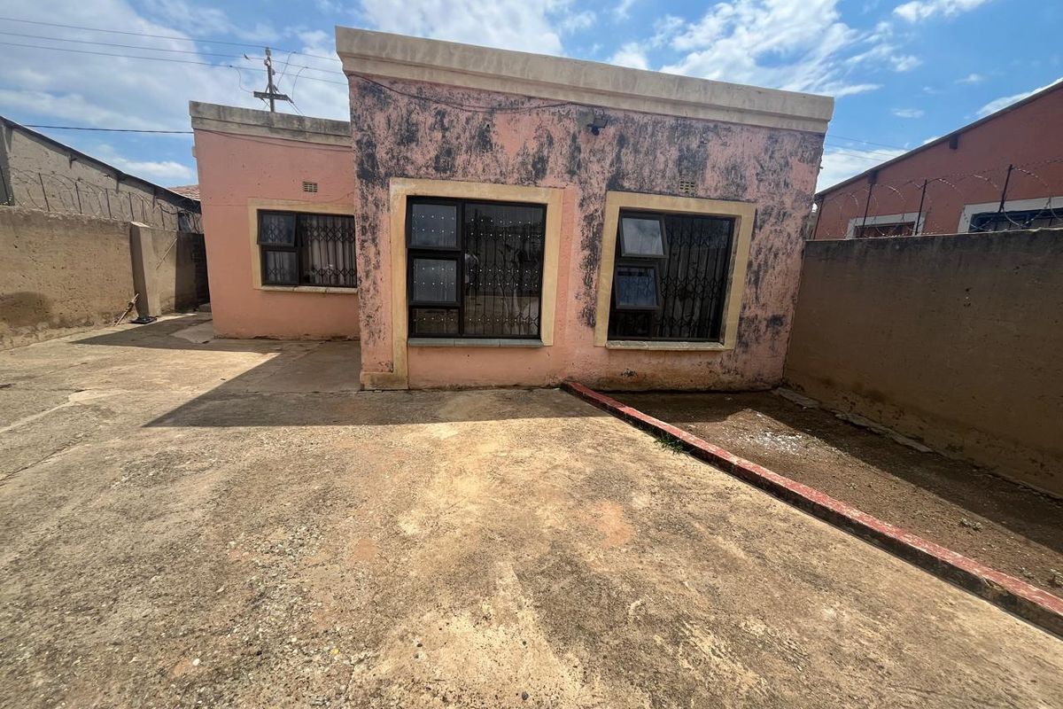 Rear exterior view of the property featuring a paved yard, boundary walls with security fencing, and a building facade requiring maintenance and repainting.