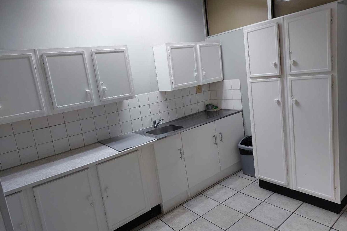 A functional office kitchenette featuring white built-in cabinetry, a stainless steel sink, tiled backsplash, and durable ceramic floor tiles.